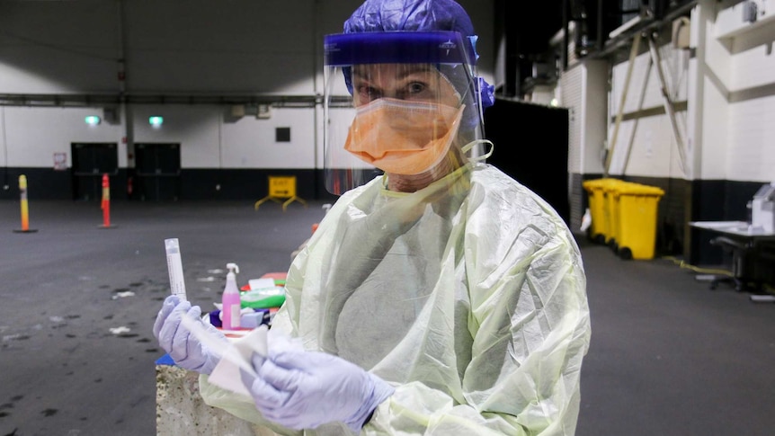 Cheryl holds a swab, wearing a protective robe, mask, face shield, gloves and plastic cap.