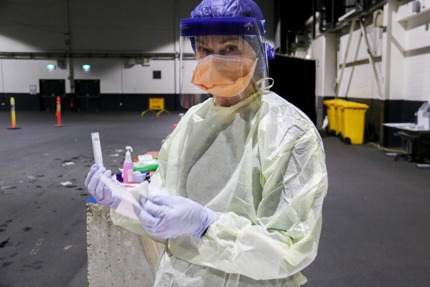 Cheryl holds a swab, wearing a protective robe, mask, face shield, gloves and plastic cap.