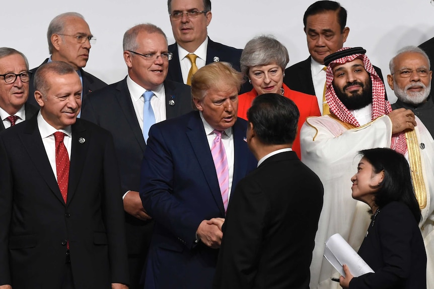 A group of leaders stand in front of a white backdrop on a stage as Donald Trump shakes hand with Xi Jinping.