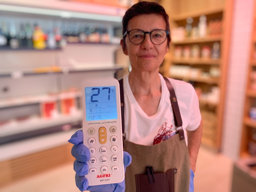 A woman wearing an apron and blue gloves holds up a thermometer displaying 27 degrees Celsius