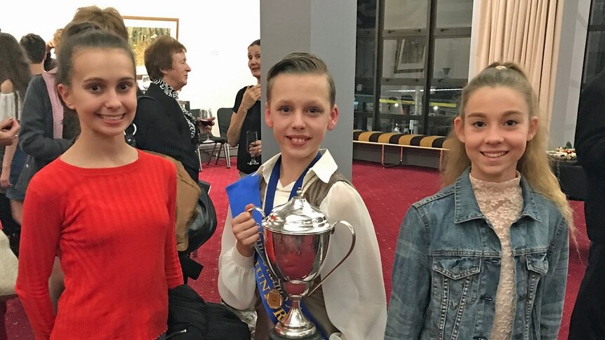 Three teenagers standing side by side smiling, with middle boy holding a large silver cup.