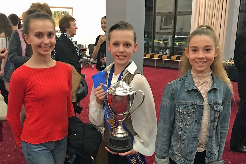 Three teenagers standing side by side smiling, with middle boy holding a large silver cup.