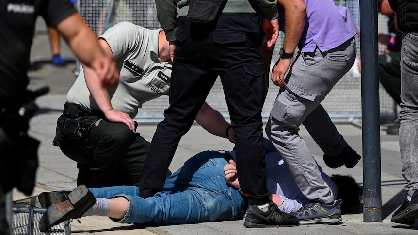 A man wearing jeans is held on the ground by police and bystanders