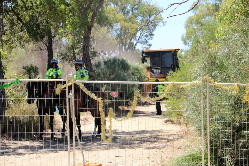 Machinery and police officers on horseback in bushland behind a fence.