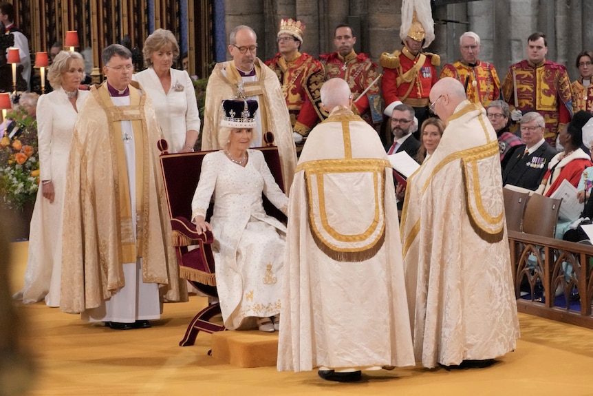 Camilla with a crown on her head sitting in a chair, surrounded by people in white robes. 