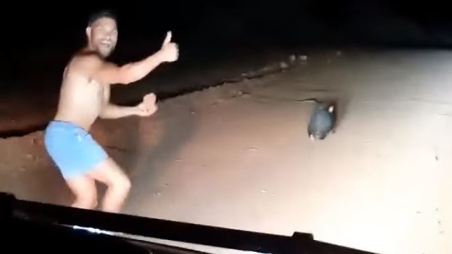 A man runs shirtless in front of a car, giving a thumbs up.