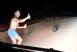 A man runs shirtless in front of a car, giving a thumbs up.