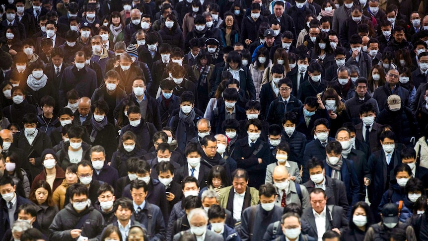 A group of Japanese people walking through a crowd with many wearing face masks