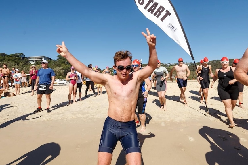 Bobby Pate, holding up his arms make V sign with hands, competes at a ocean swimming race on a beach at a triathlon event.