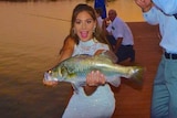 A woman in a wedding dress is excited to hold a big barramundi as a man reels in over her head. Sunset.