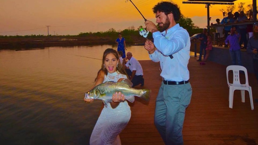 A woman in a wedding dress is excited to hold a big barramundi as a man reels in over her head. Sunset.