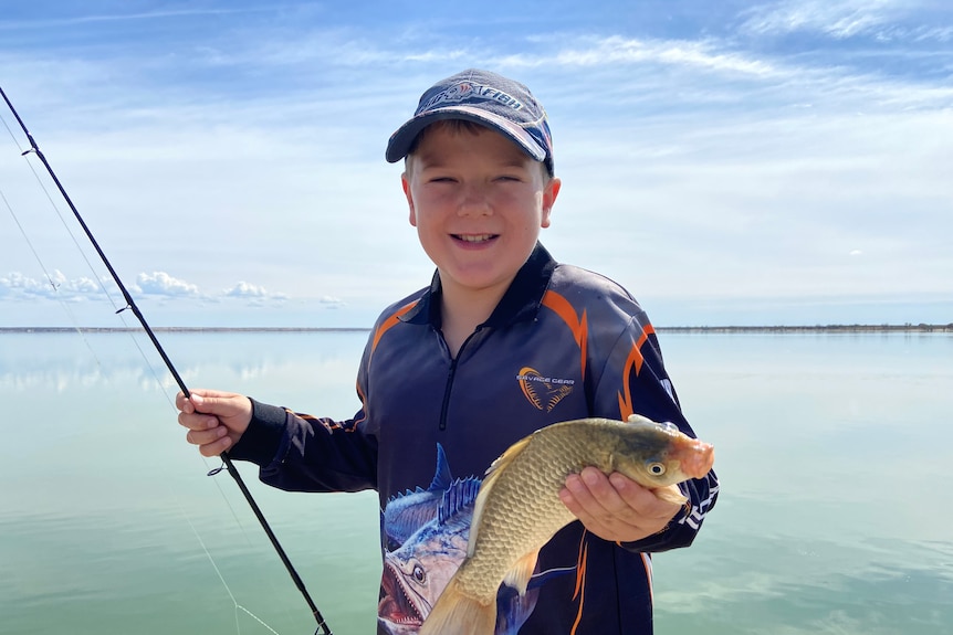 A boy wearing a long-sleeved blue fishing top holds a carp with blue water in background.