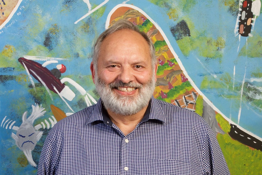 A man white grey hair and beard stands in front of a mural.