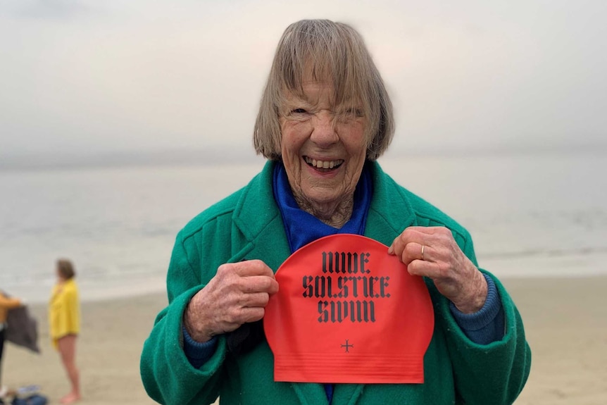 An elderly woman holds up a red swimming cap on a beach at dawn.