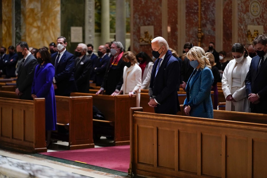 Joe and Jill Biden wearing masks and with heads bowed stand in a church pew along with many other people in a church.