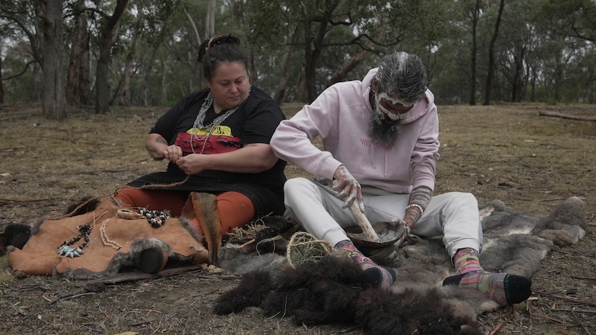 An Aboriginal man sits on the ground with white cultural pigment on his face. An Aboriginal woman is seated next to him.