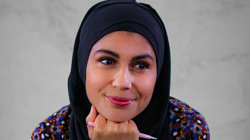 A woman wearing a hijab and colourful shirt holding a pencil with her hand under her chin.