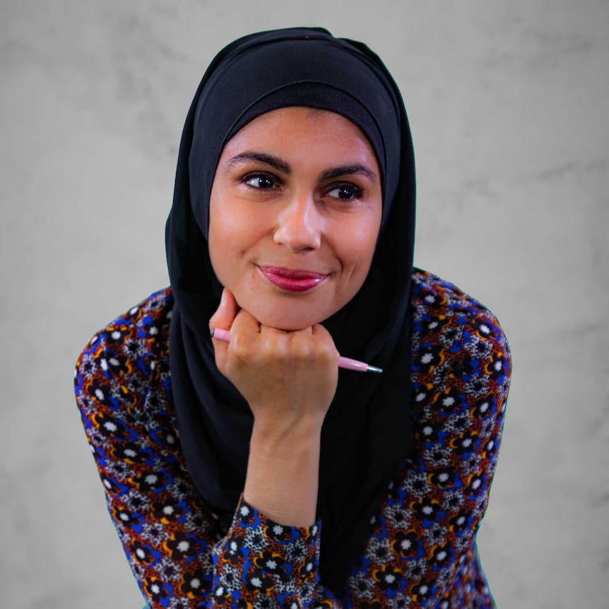 A woman wearing a hijab and colourful shirt holding a pencil with her hand under her chin.