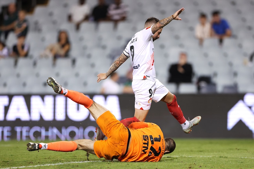 A goalkeeper lies on the ground with the ball (hidden) as an opposition striker swings his leg through after failing to connect.