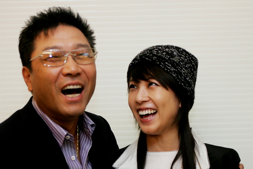 A man in a purple shirt and dark suit, with transparent sunglasses, smiles with his arm around a young woman