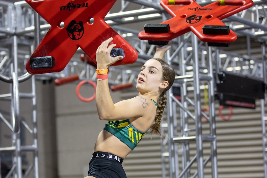 A teenage girl in sports gear concentrates as she grasps and swings between two "X"-shaped plates hanging from the ceiling.