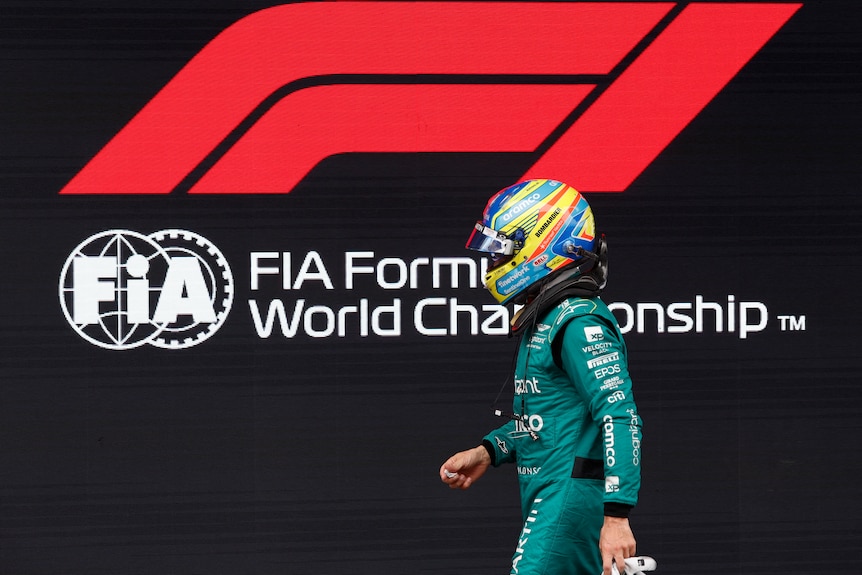 Fernando Alonso, wearing his green race suit and helmet, walks past a Formula 1 logo sign, at a race track.