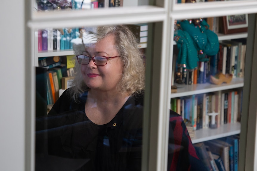 Woman wearing red glasses sits behind window at her desk.