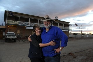 A woman in a dark shirt hugs her smiling, bearded, hat-wearing husband outside their country pub as the sun sets behind them.