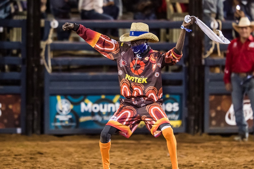 An Indigenous man wearing brown and orange clothes and a cowboy hat dances in a rodeo arena