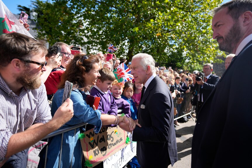 An elderly man in a dark suit shakes a woman's hand as a crowd of onlookers waits to see him.
