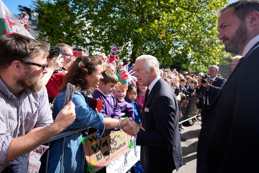 An elderly man in a dark suit shakes a woman's hand as a crowd of onlookers waits to see him.