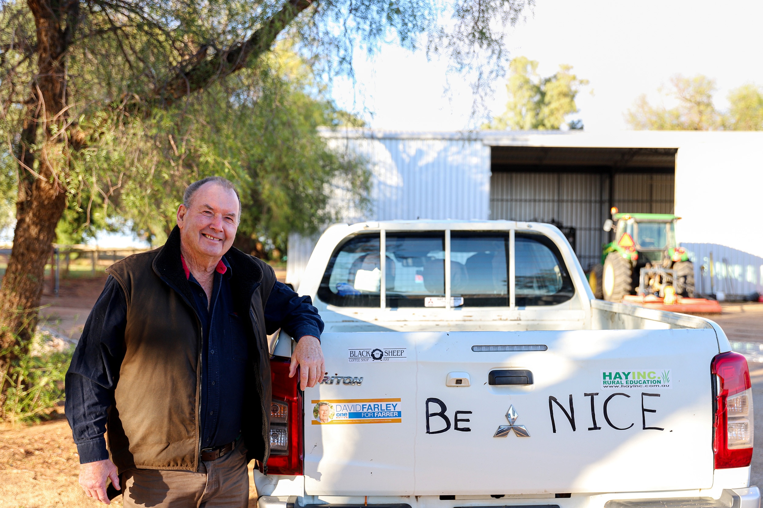 A man wearing a blue shirt and a brown jacket leans on a white utility vehicle that has writing saying 'Be Nice'