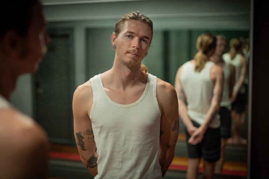 Young man wearing white singlet stands with hands behind back, in front of mirror.