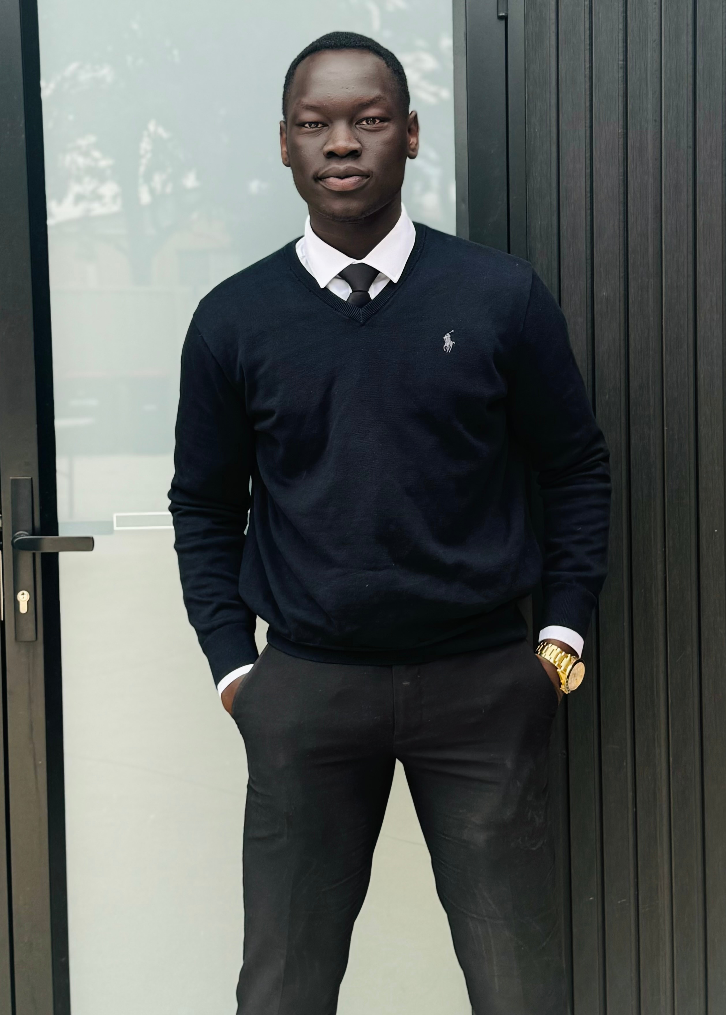 A young Black man wear a white shirt, black tie and blue jumper, and stands looking thoughtful.