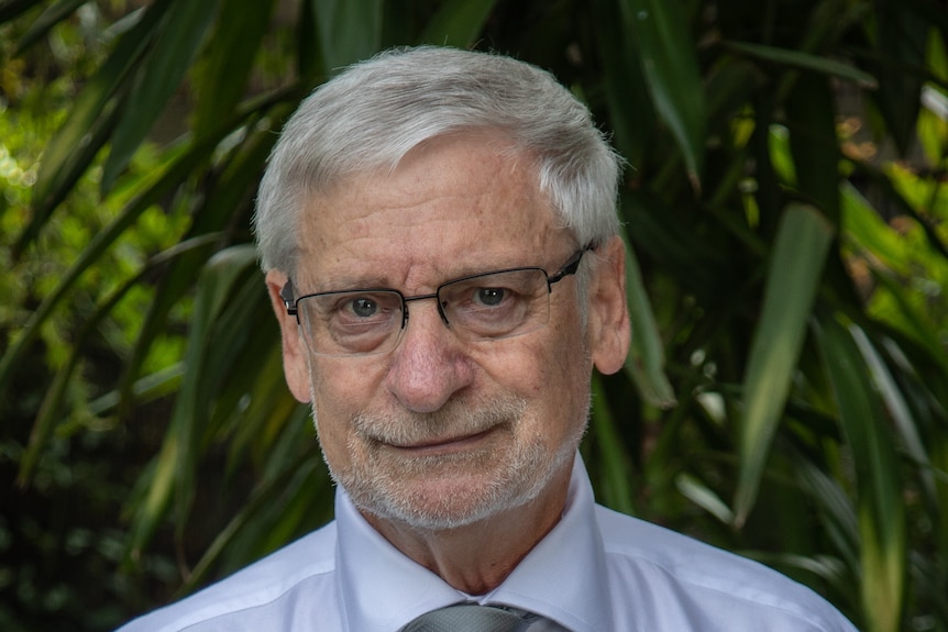 An older man with white hair and a beard and glasses stands in front of plants.