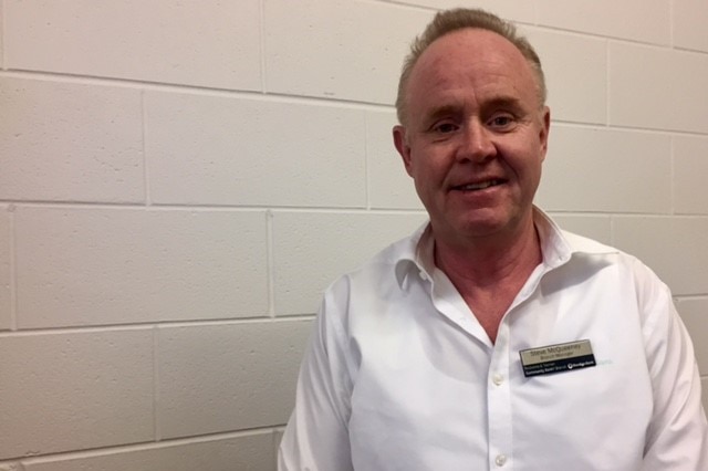 Balding man with white business shirt and name tag stands in front of brick wall