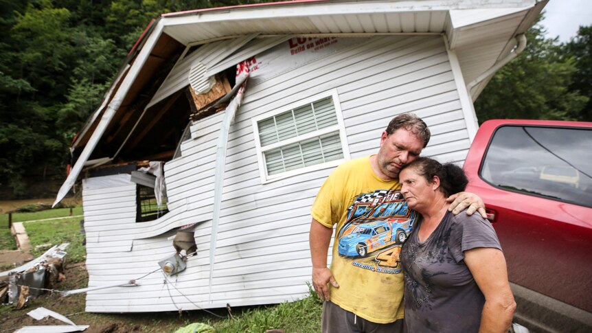 Man and woman wrap their arms over each other to comfort each other in front of flood damaged home.