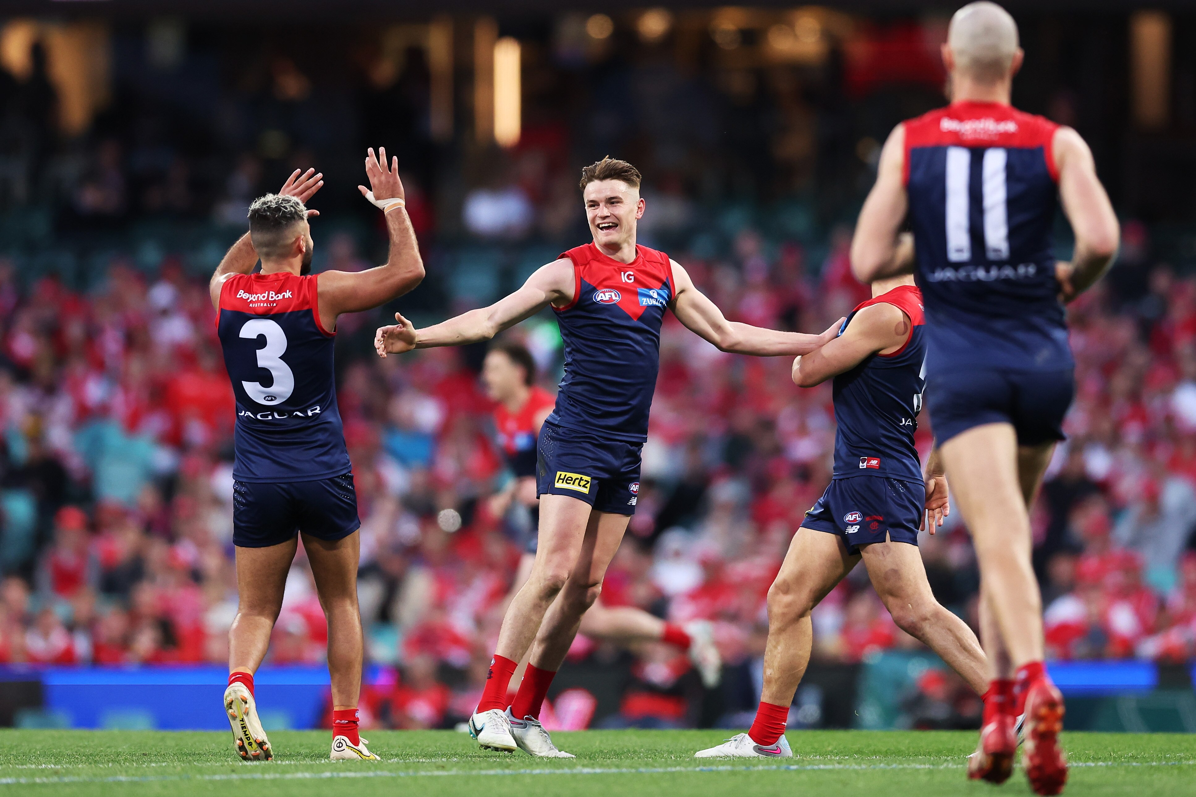 A Melbourne AFL player with his head wrapped in a bandage stands with arms up in celebration after a goal.