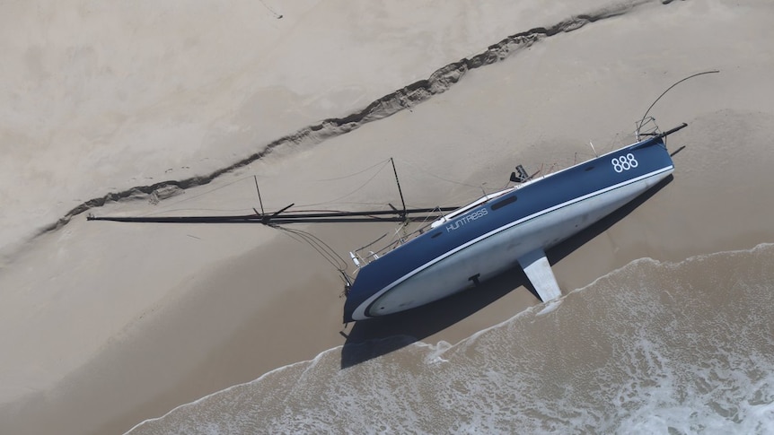 A yacht lies on its side on a beach with its mast askew.