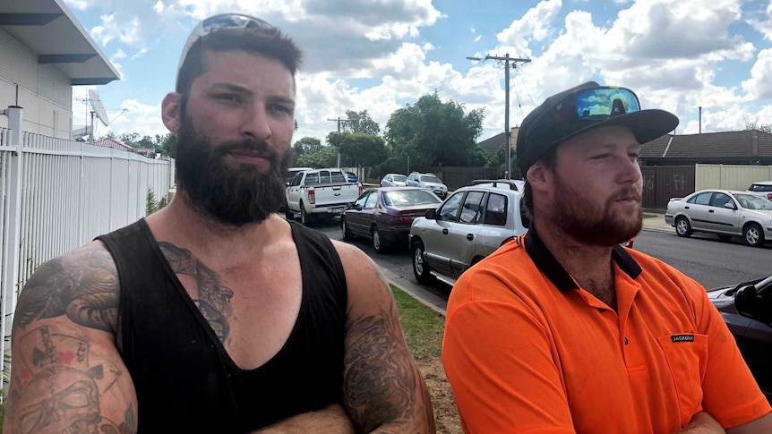 Two men who say they are owed money stand with arms folded outside a house.