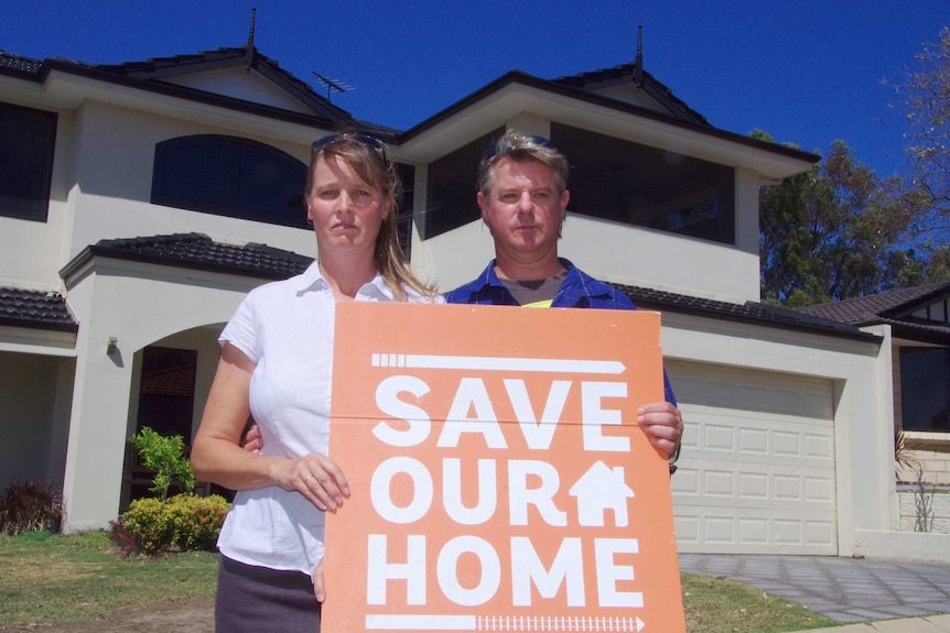 Tanya and Damon Smirke stand in front of their home holding a Save Our Home sign.