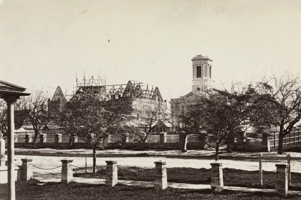 Black and white photo showing tree lined street with half constructed building and church behind the trees