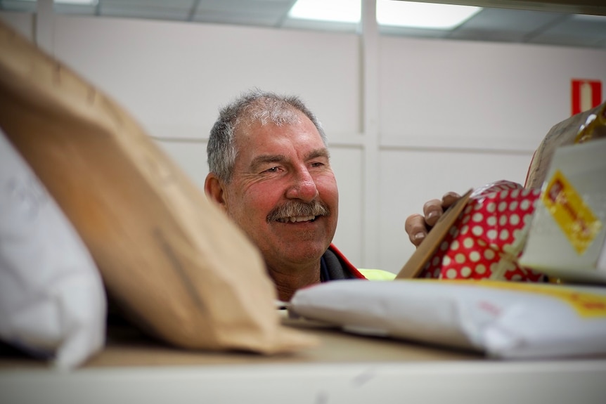 A man with thinning grey hair and a thick moustache sorts through a pile of mail. He is smiling.