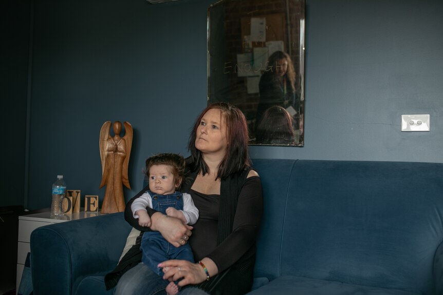 A woman sits on a couch with a baby on her lap. Another woman can be seen reflected in a mirror on the wall.