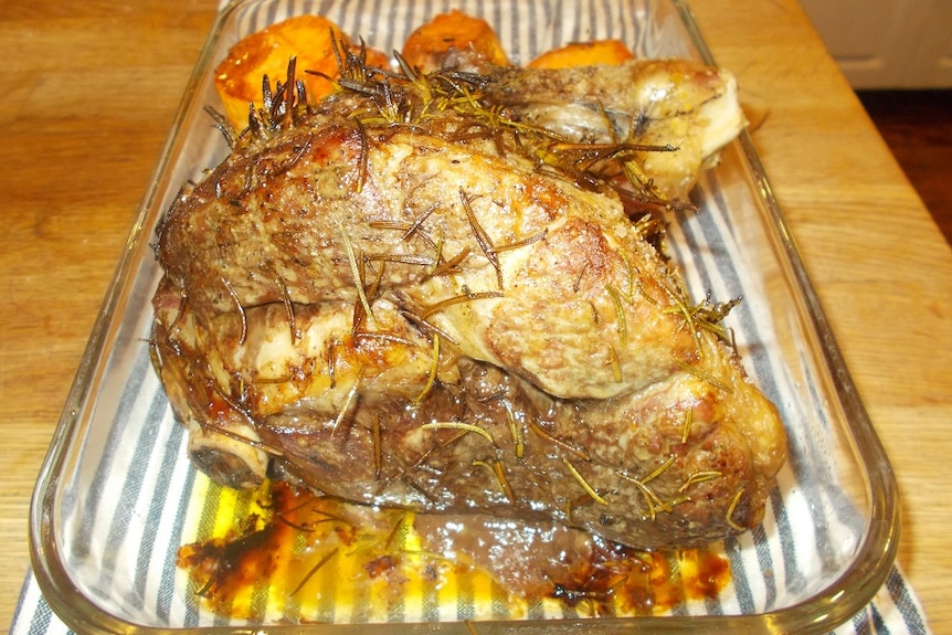 A cooked lamb roast in a clear dish with potatoes on a kitchen bench.