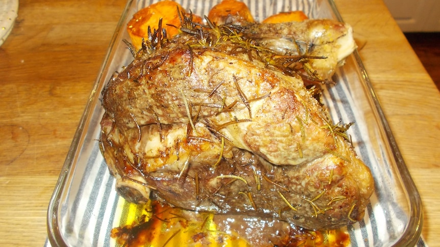 A cooked lamb roast in a clear dish with potatoes on a kitchen bench.