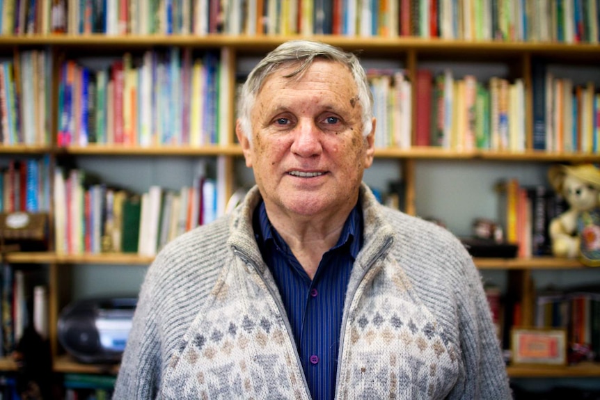 John Marsden stands in front of a bookshelf as he smiles at the camera.