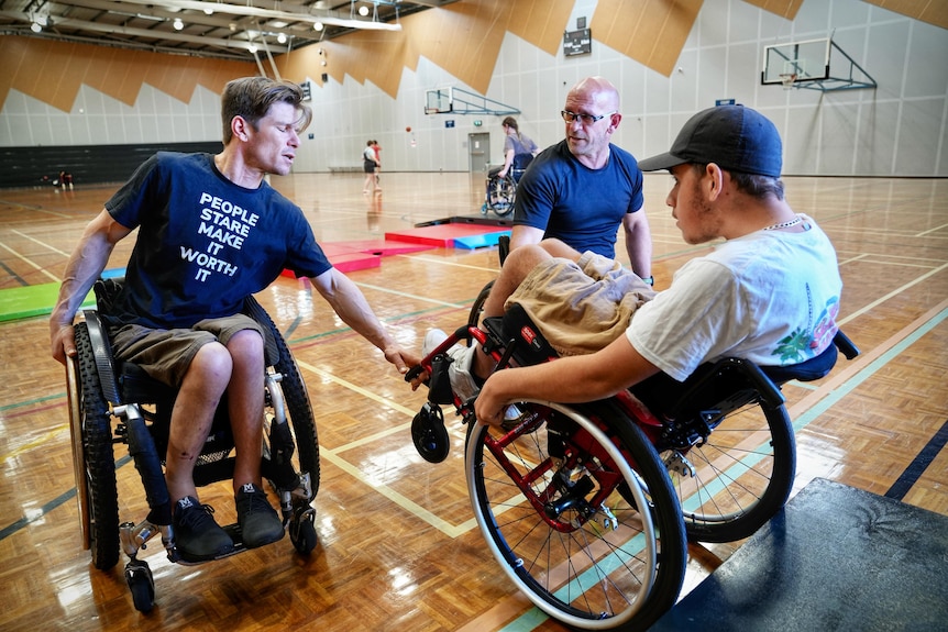 Three men in wheelchairs, with one of them helping to tip up another into a wheelie position.