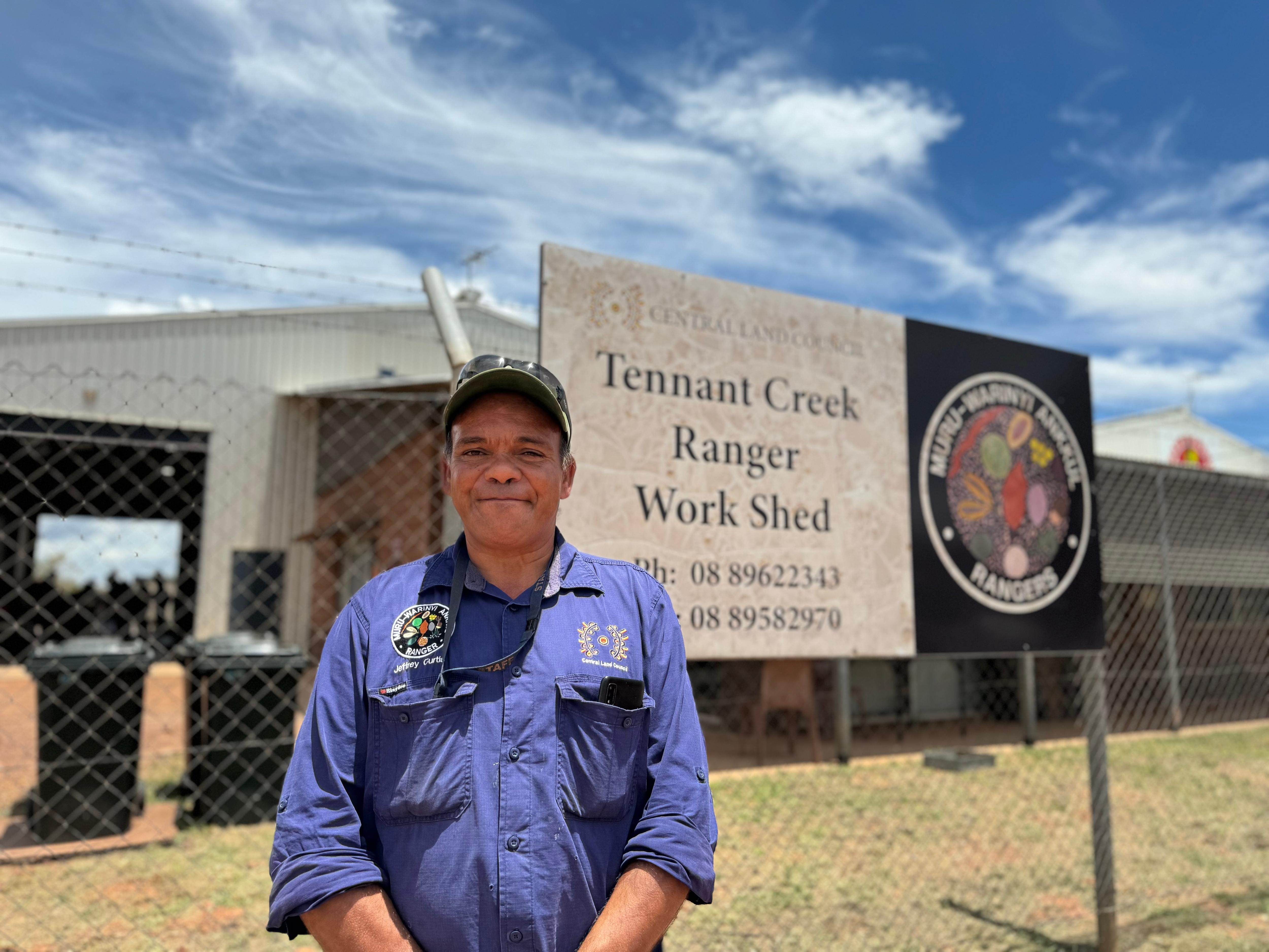 An Aboriginal man wearing a cap and blue work shirt looks at the camera in front a Tennant Creek Ranger Workshed sign.