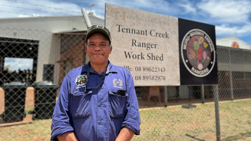 An Aboriginal man wearing a cap and blue work shirt looks at the camera in front a Tennant Creek Ranger Workshed sign.